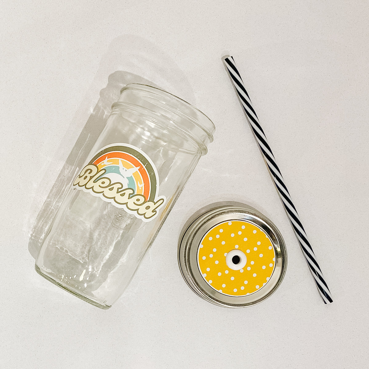 Flat lay of a mason jar tumbler that says &quot;blessed&quot;, a yellow straw lid with white polka dots, and a black and white striped straw. 