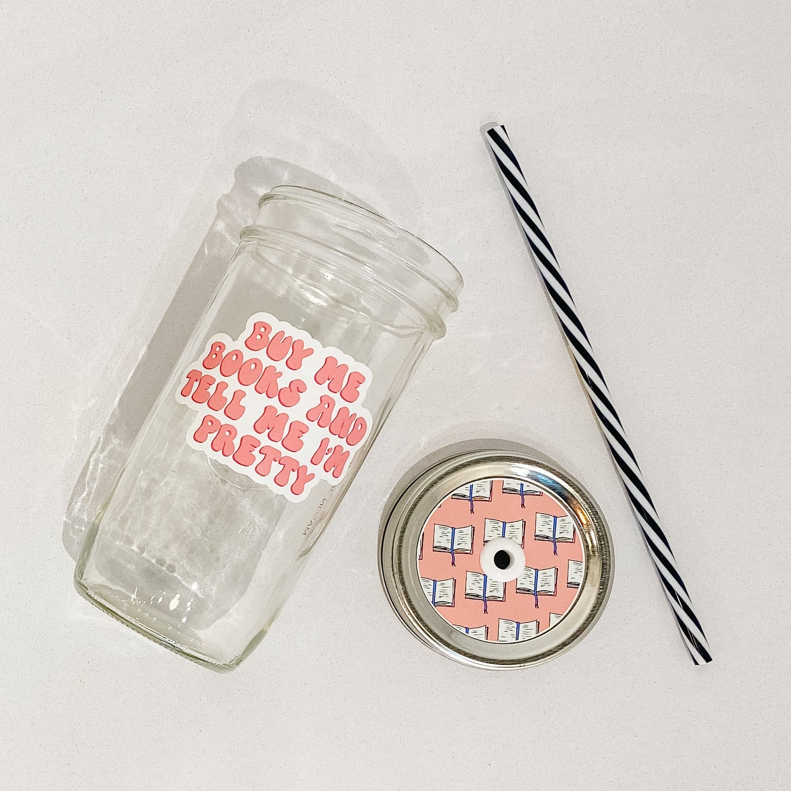 Flat lay of a mason jar tumbler that says “buy me books and tell me I’m pretty”, a pink straw lid with a repeating pattern of an opened book, and a black and white striped straw. 