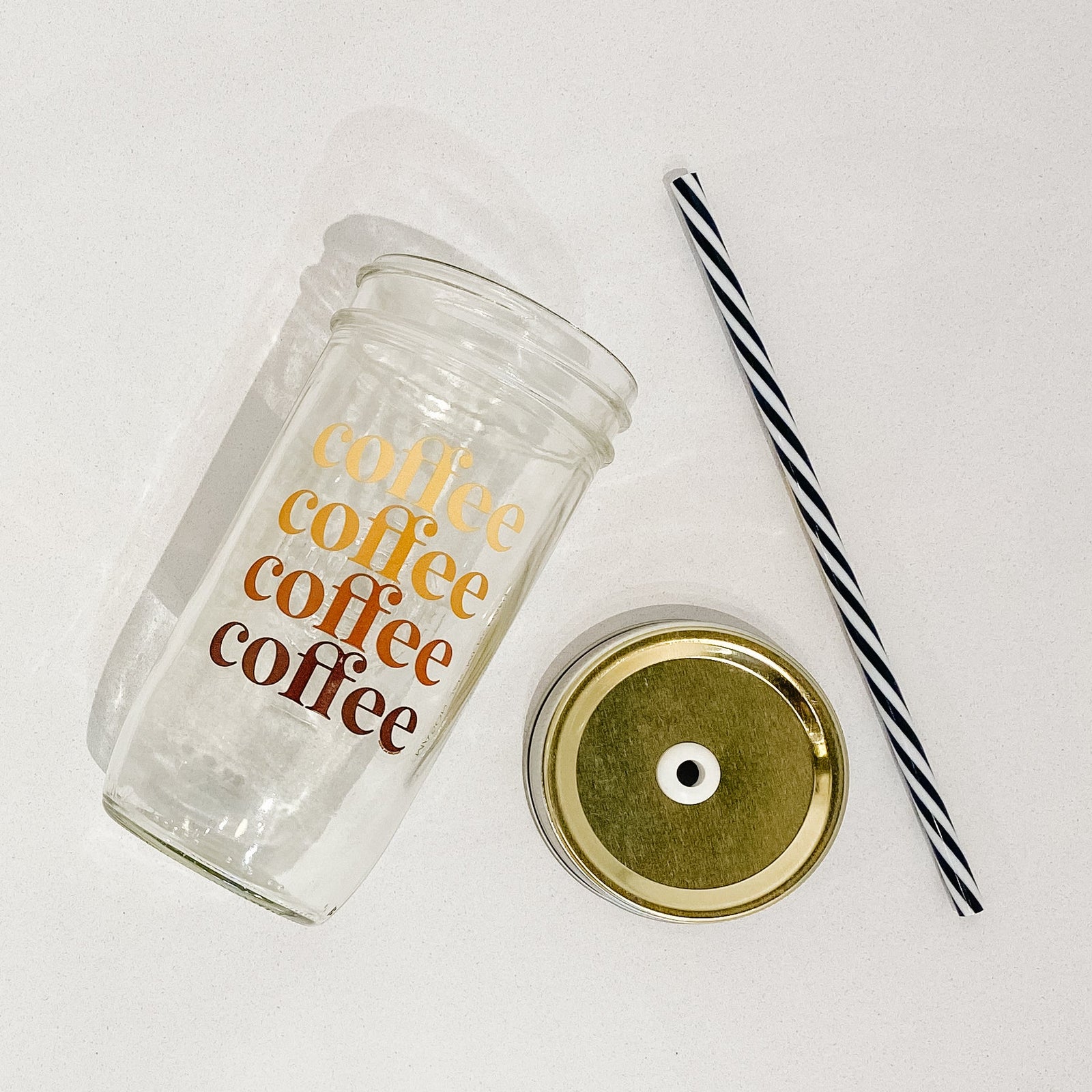 Flat lay of a mason jar tumbler that says “coffee” 4 times in different shades of brown, a straw lid in gold, and a black and white striped straw.