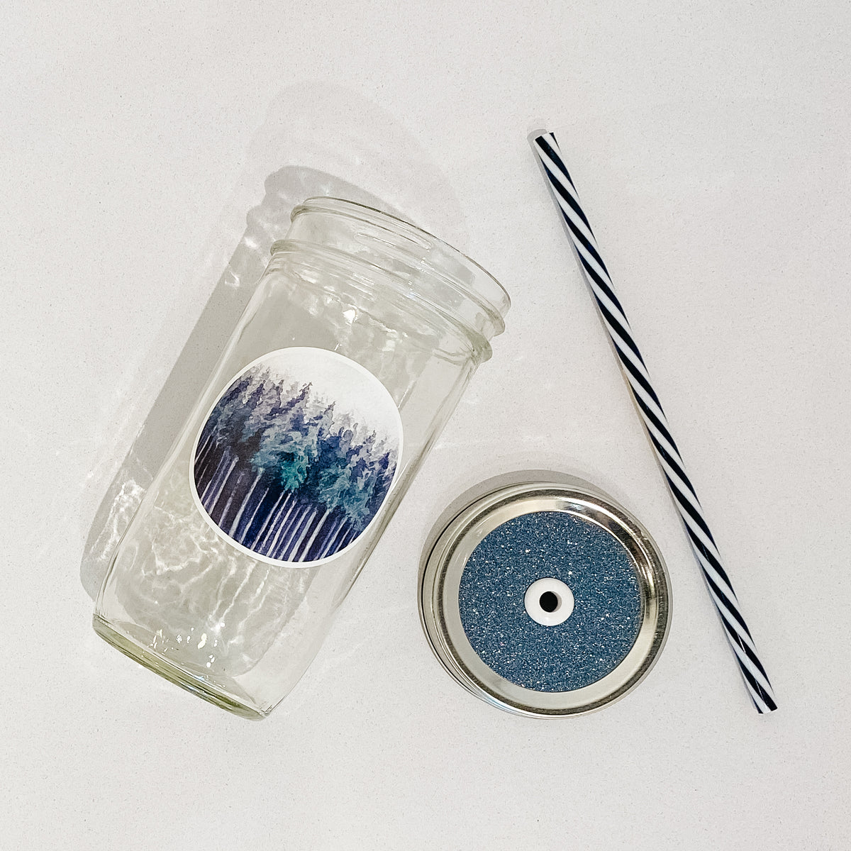 Flat lay of a mason jar tumbler that has a decal of dark trees, a dark grey glitter lid, and black and white striped straw. 