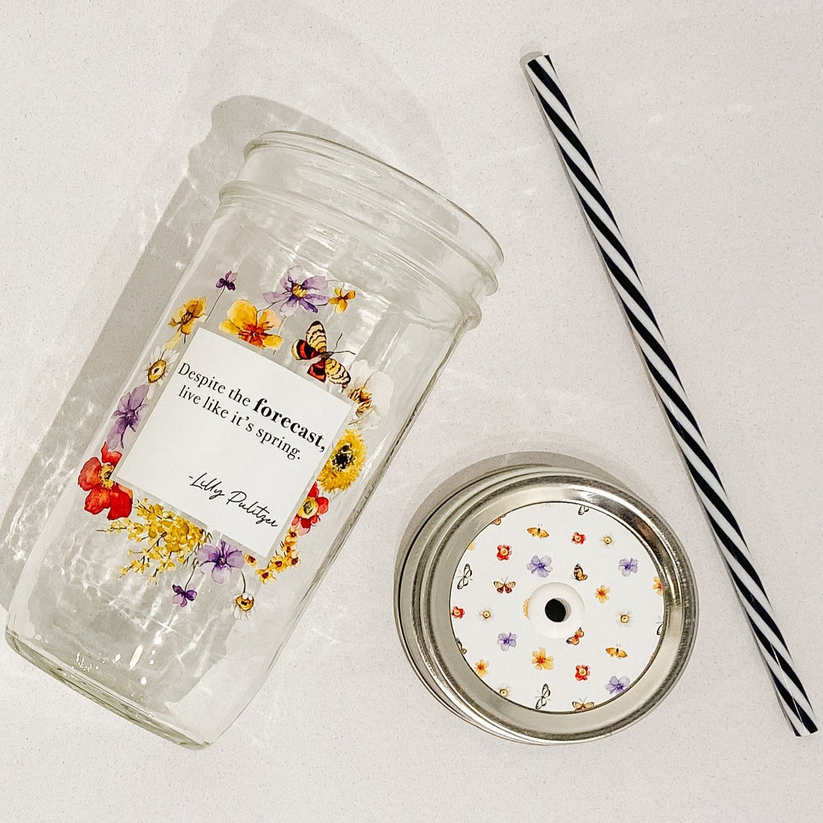 Flat lay of a mason jar tumbler that says “Despite the forecast, live like it’s spring. -Lilly Pulitzer”, a straw lid that is white with florals and butterflies creating a repeating pattern, and a black and white striped straw