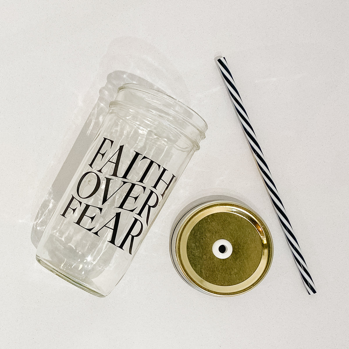 Flat lay of a mason jar tumbler that says “faith over fear”, a gold straw lid, and a black and white striped straw.