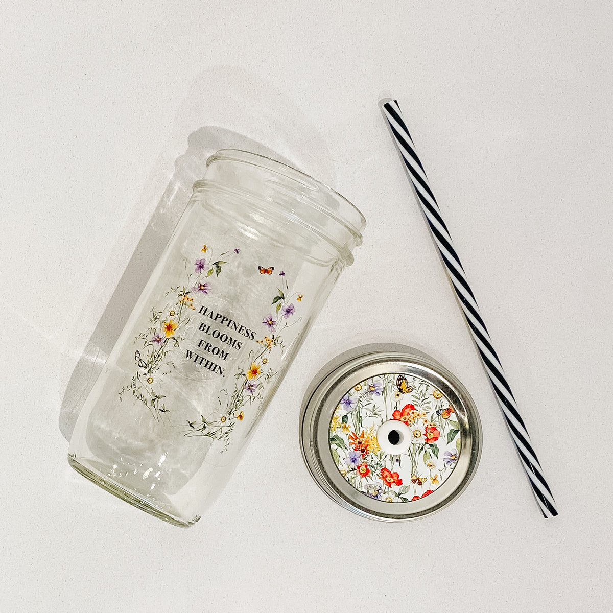 Flat lay of a mason jar tumbler that says “happiness blooms from within”, a straw lid that is busy with different flowers, greenery, and butterflies, and a black and white straw. 