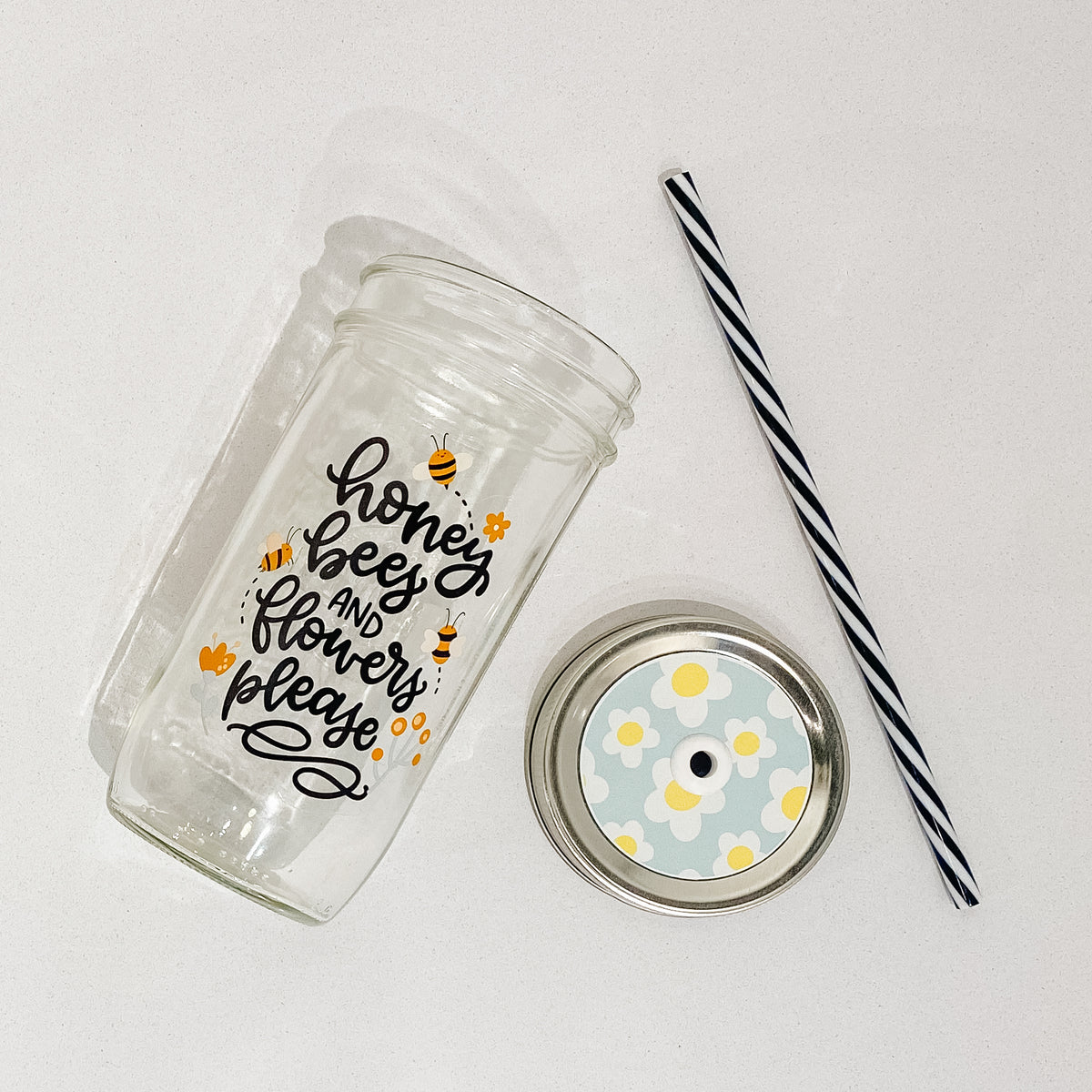 Flat lay of a mason jar tumbler that says “honey, bees, and flowers please”, a straw lid that has a light blue background and white flowers with yellow centres, and a black and white striped straw. 