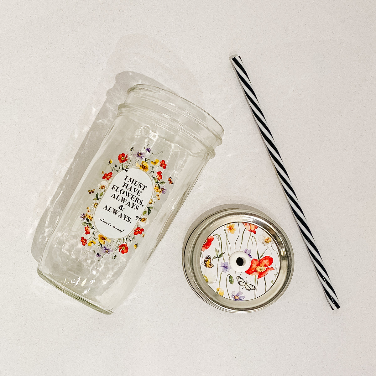 Flat lay of a mason jar tumbler that says &quot;I must have flowers, always &amp; always. -Claude Monet”, a white straw lid with florals and butterflies, and a black and white striped straw. 