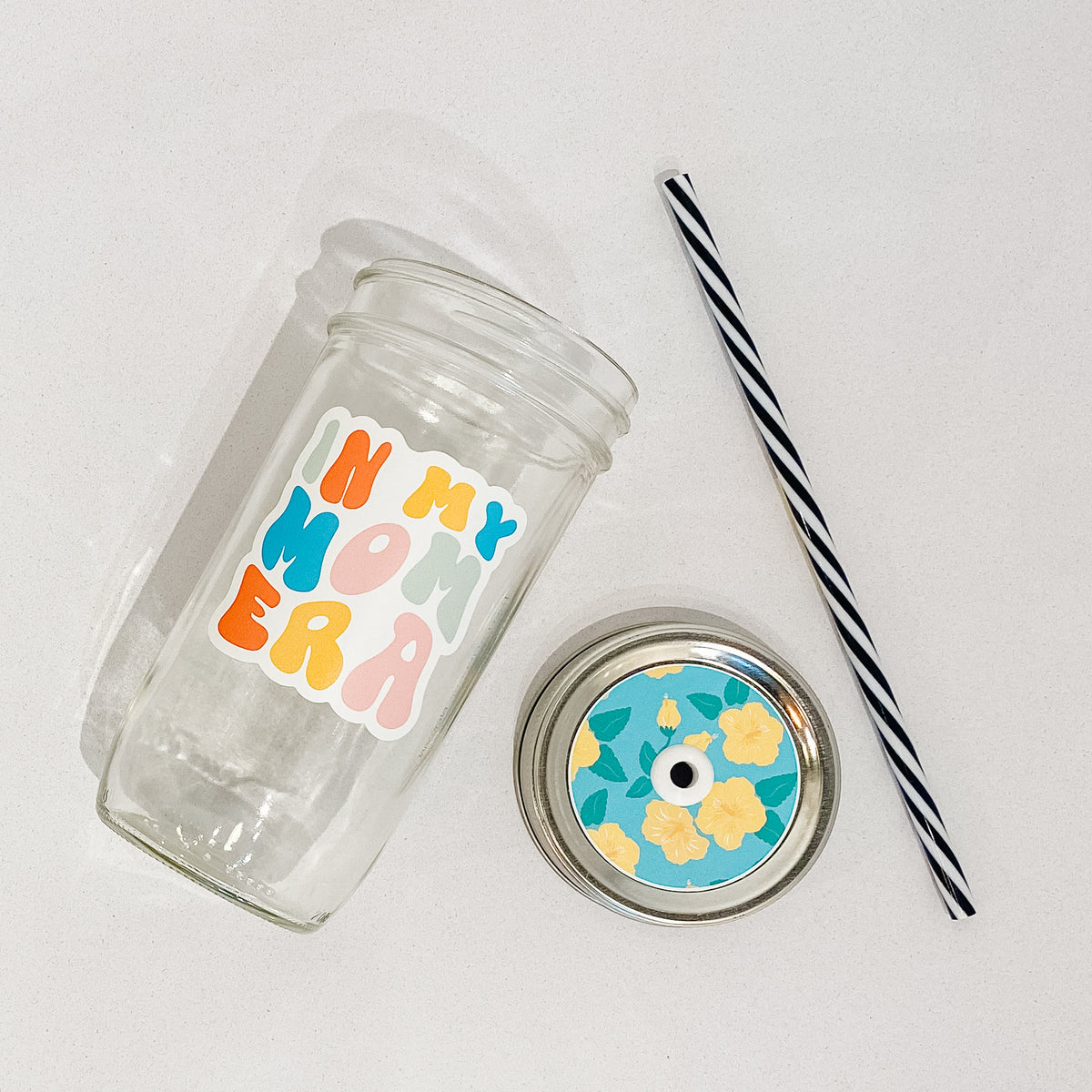 Flat lay of a mason jar tumbler that says &quot;in my mom era&quot;, a straw lid of yellow flowers on a blue background, and a black and white striped straw. 