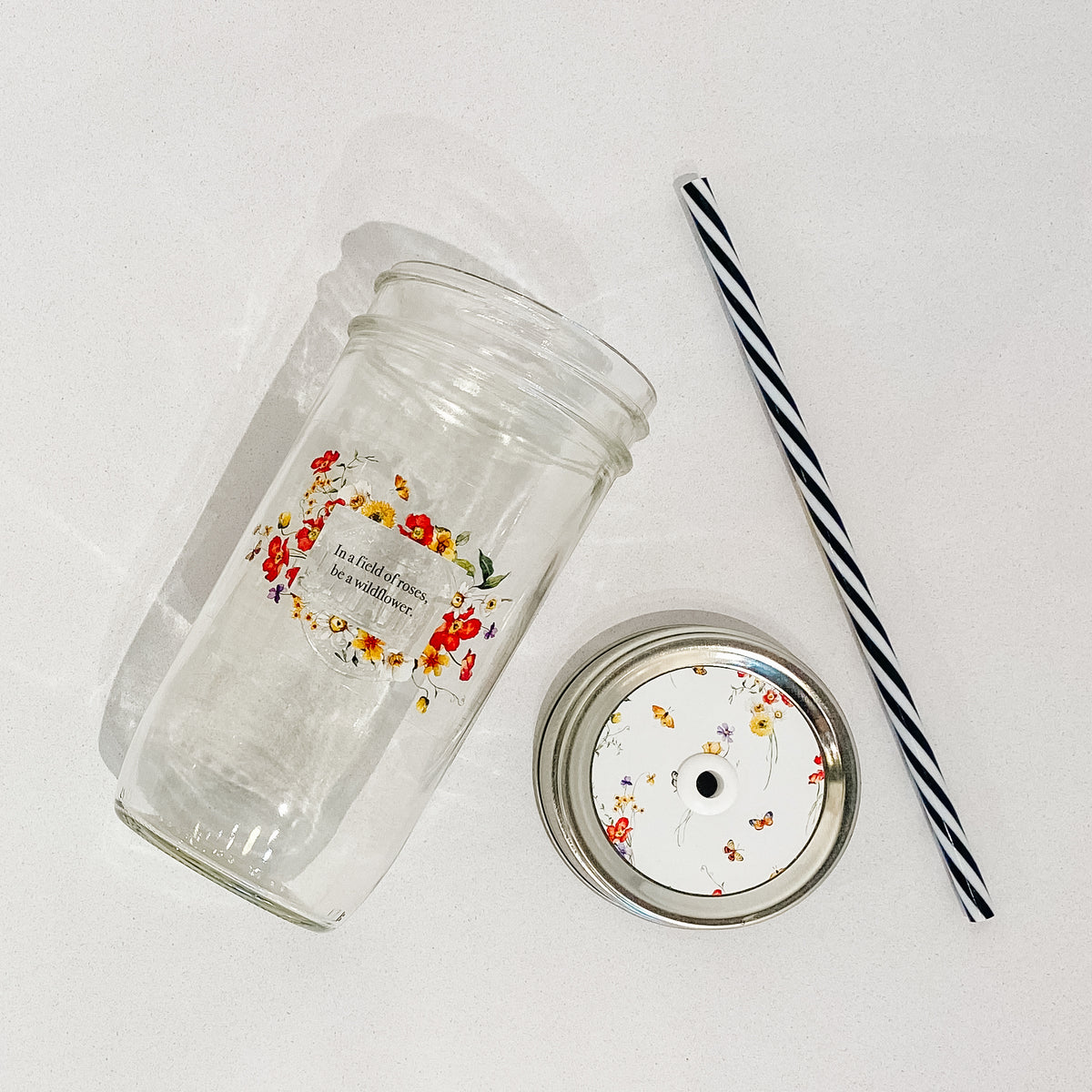 Flat lay of a mason jar tumbler that says &quot;in a field of roses, be a wildflower”, a straw lid that is white with different flowers and butterflies spread out on it, and a black and white striped straw.