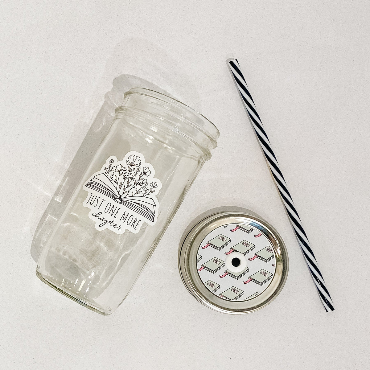 Flat lay of a mason jar tumbler that says “just one more chapter” with flowers growing out of an open book, a straw lid that has a repeating pattern of a green book, and a black and white striped straw.