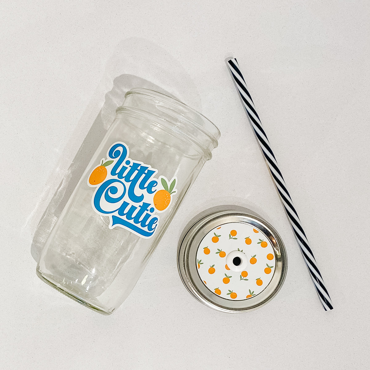 Flat lay of a mason jar tumbler that says “little cutie”, a straw lid that has small oranges (fruit) on a white background, and a black and white striped straw. 