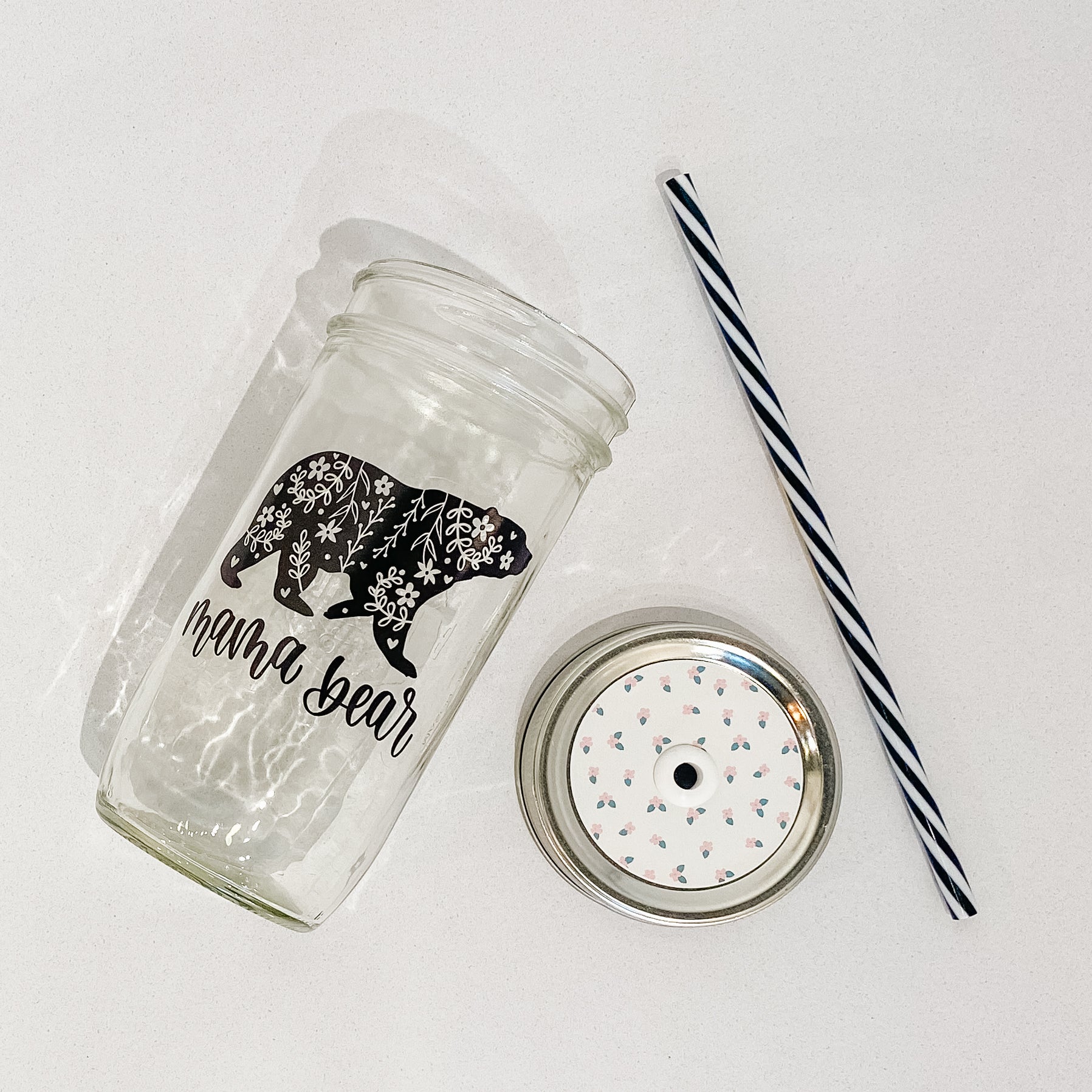 Mason jar tumbler filled with a citrus water drink and has a gold lid and straw and a calligraphy print that reads "mama bear" with white floral patterns in a black bear graphic. Photographed against a marble countertop.