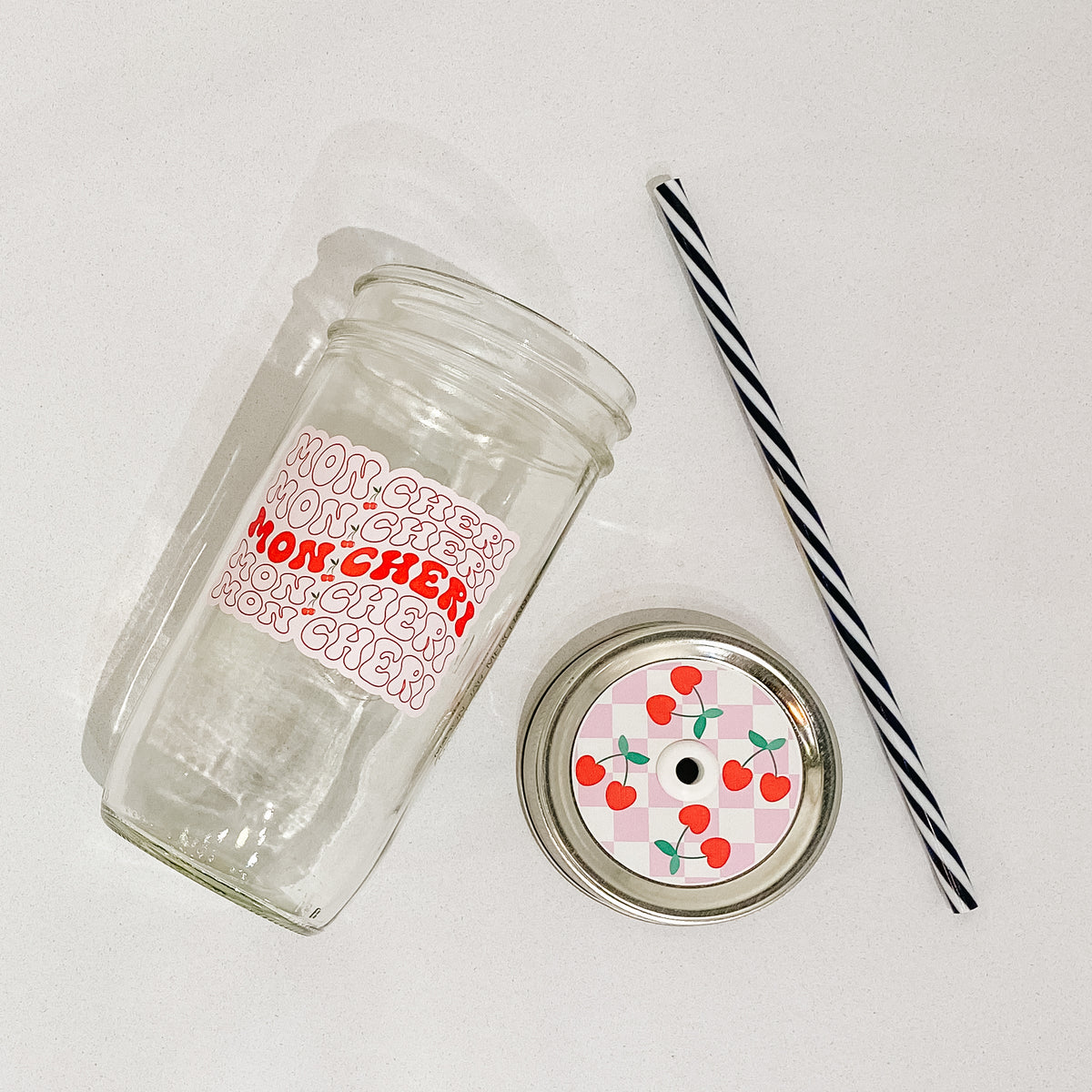 Flat lay of a mason jar tumbler that says “mon cheri”, a straw lid with 4 cherries (one in each quadrant) on a pink and white checkered background, and a black and white straw.