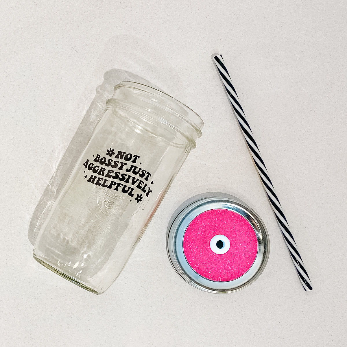 Flat lay of a mason jar tumbler that says “not bossy just aggressively helpful”, a hot pink glitter straw lid, and a black and white striped straw. 