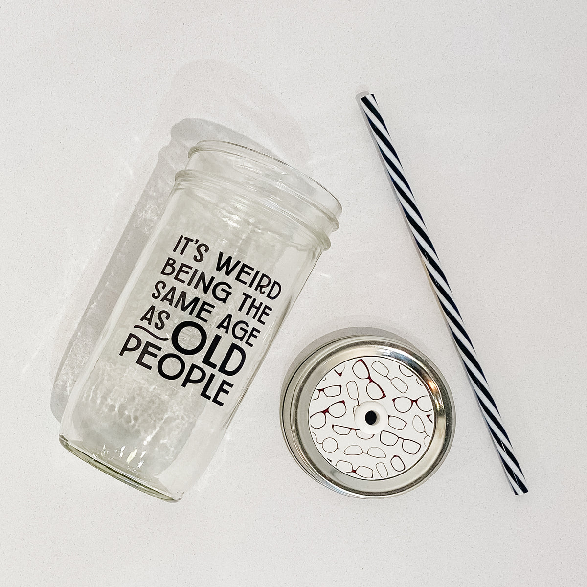 Flat lay of a mason jar tumbler that says “It’s weird being the same age as old people”, a straw lid that has different pairs of glasses on it, and a black and white straw. 