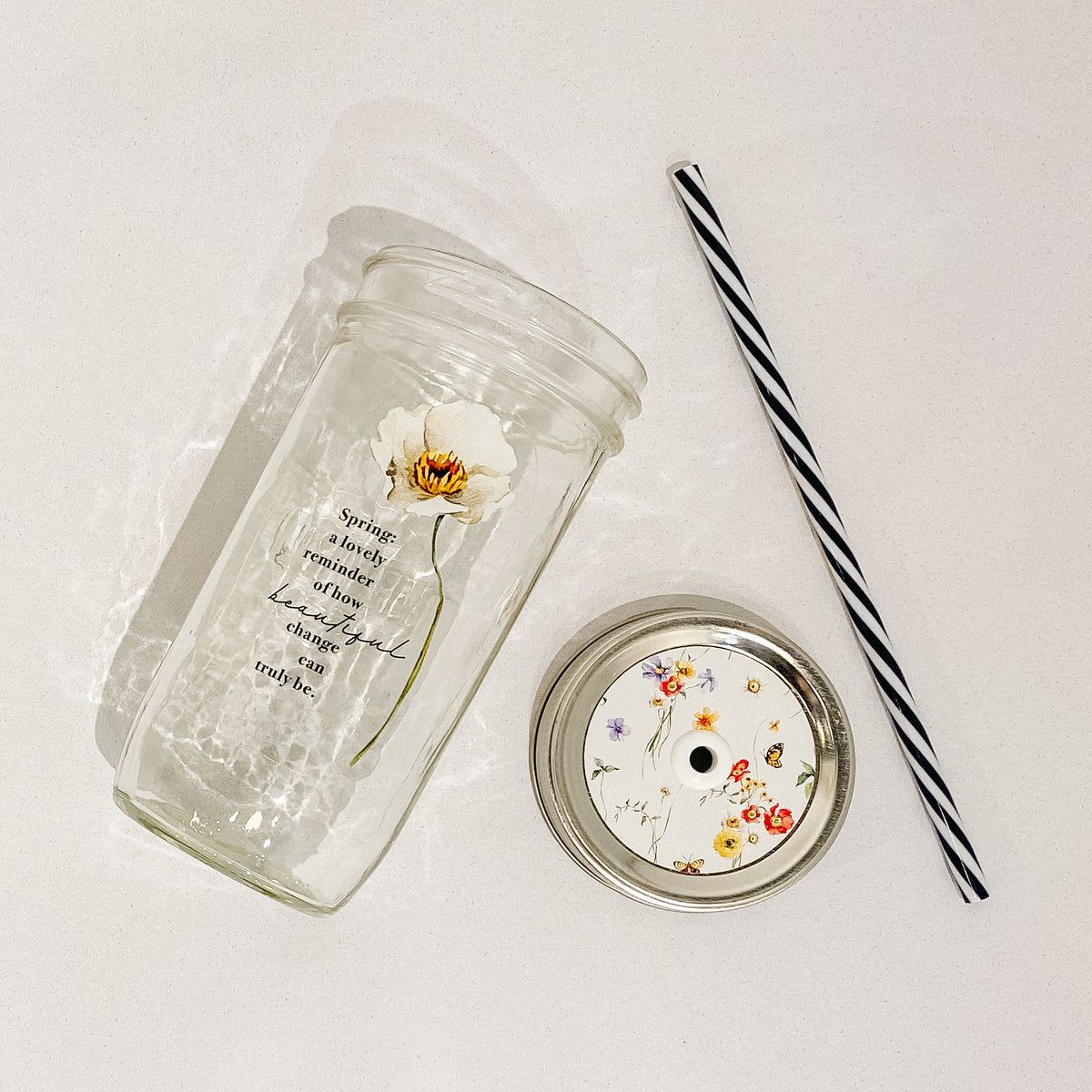 Flat lay of a mason jar tumbler that says “Spring: a lovely reminder of how beautiful change can truly be.”, a straw lid that is green with different flowers and butterflies spread across it, and a black and white striped straw.