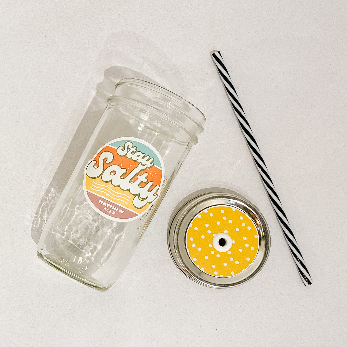 Flat lay of a mason jar tumbler that says &quot;stay salty&quot;. a yellow straw lid with white polka dots, and a black and white striped straw. 