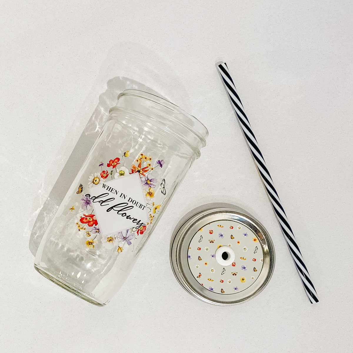 Flat lay of a mason jar tumbler that says “when in doubt add flowers”, a green straw lid with flowers and butterflies creating a repeating pattern, and a black and white striped straw. 