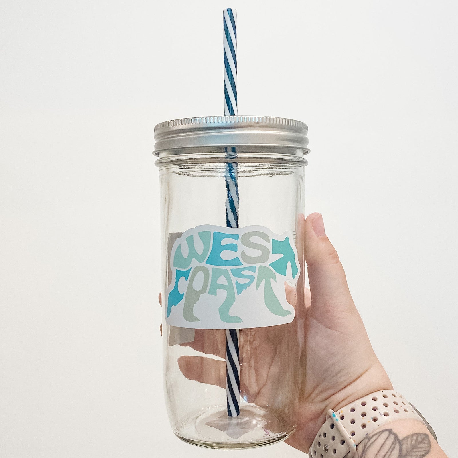 Hand holding mason jar tumbler that reads  west coast in the shape of a bear. Multiple shades of blues/greens