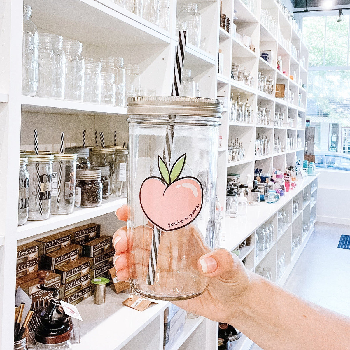 A woman holding a transparent mason jar tumbler with a cute drawing or picture of a peach. Inside the peach is a text that says: You're a peach." It has a black and white straw and behind it is a white cabinet or a storage full of different types of mason jar tumblers.