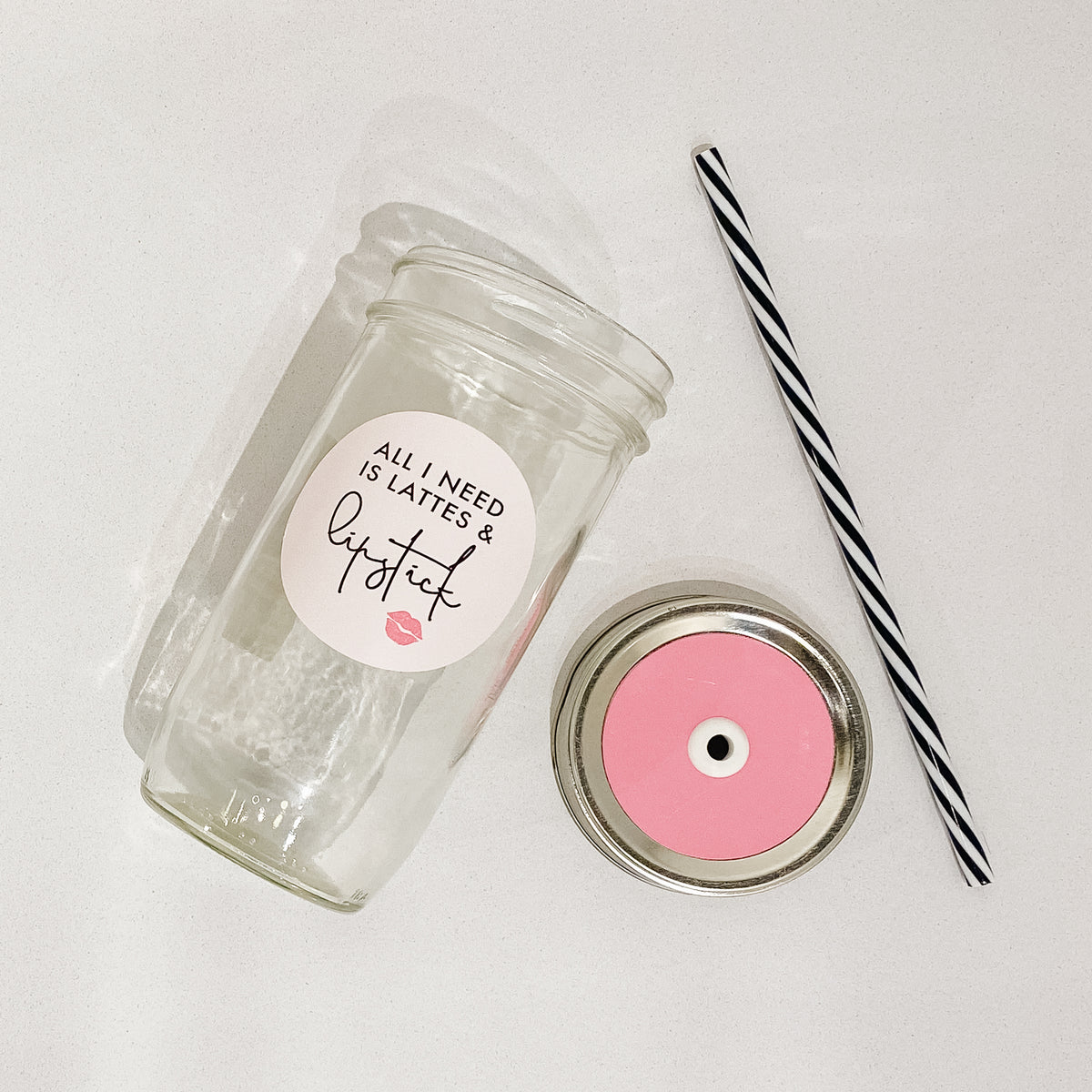 Flat lay of a mason jar tumbler that says “all I need is lattes &amp; lipstick”, a straw lid that is bubblegum pink, and a black and white striped straw. 