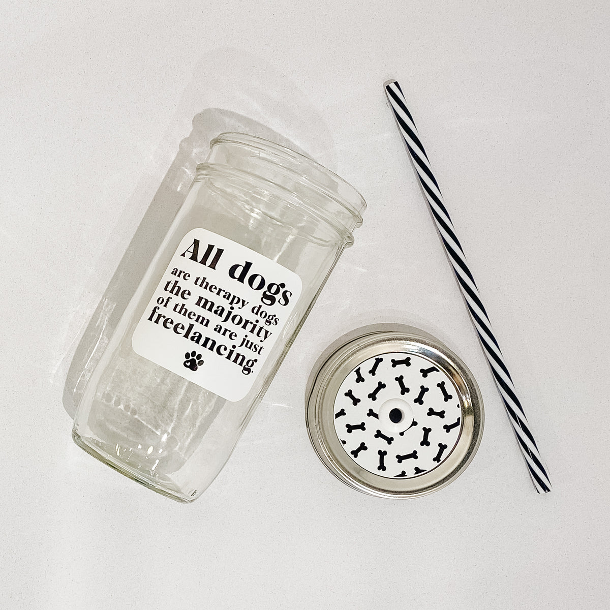 Flat lay of a mason jar tumbler that says &quot;all dogs are therapy dogs the majority of them are just freelancing”, a straw lid with black bones over it on a white background, and a black and white striped straw. 