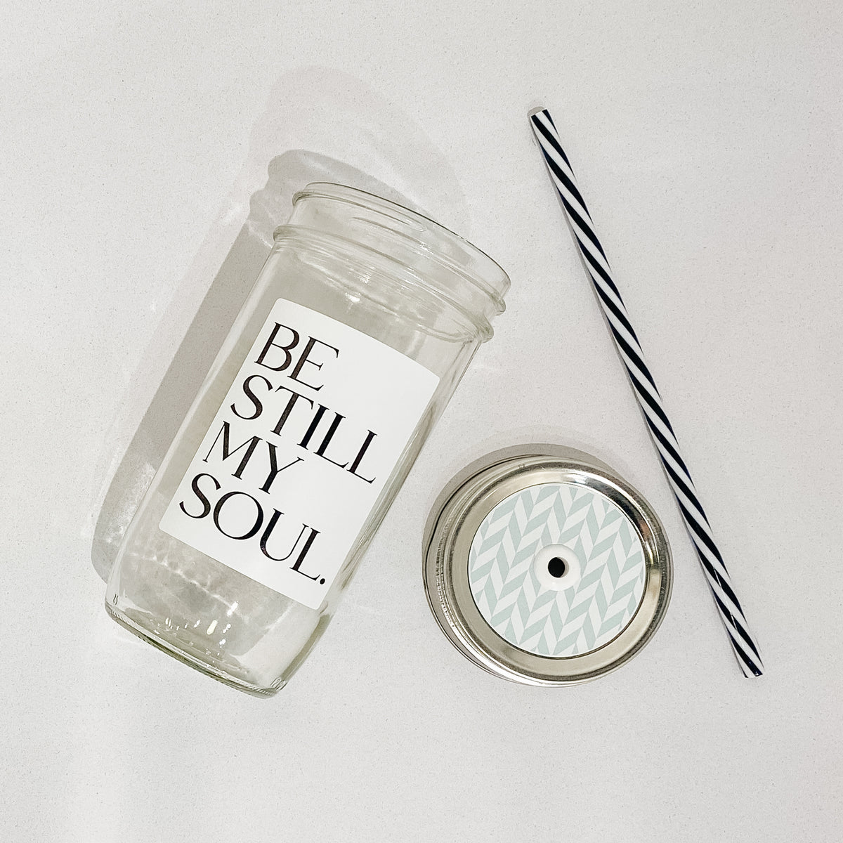 Flat lay of a mason jar tumbler that says &quot;be still my soul”, a straw lid that has a mint and white chevron-esque pattern, and a black and white striped straw.