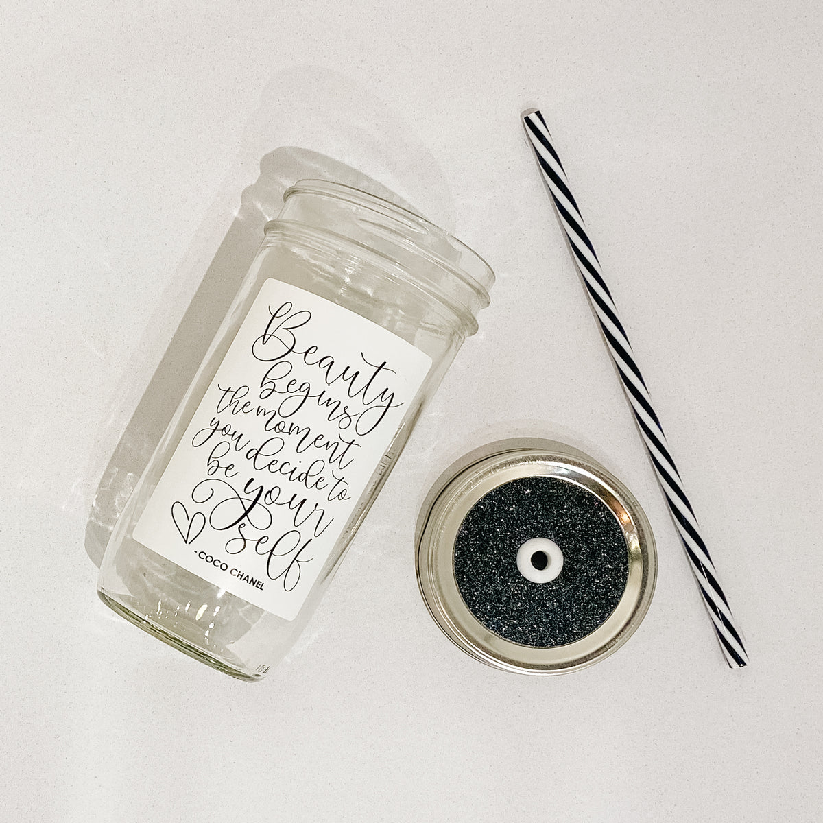 Flat lay of a mason jar tumbler that says “Beauty begins the moment you decide to be your self -Coco Chanel”, a straw lid that is black with silver glitter, and a black and white striped straw.