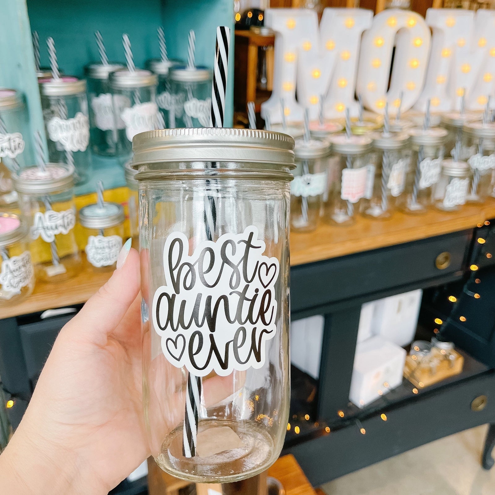 Hand holding a mason jar tumbler that says “best auntie ever” in black script on a white background. There are 2 small hearts beside the words.