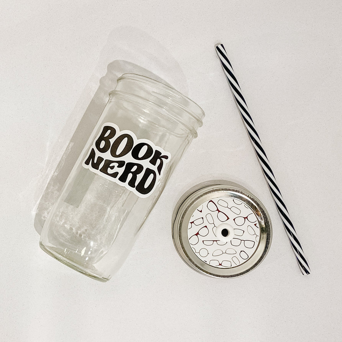 Flat lay of a mason jar tumbler that says "book nerd”, a straw lid with images of various reading glasses, and a black and white striped straw. 
