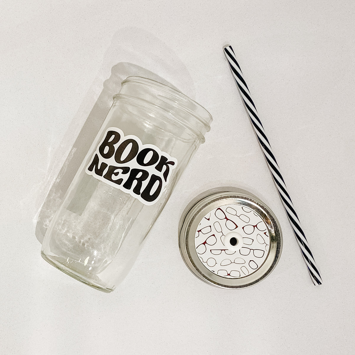 Flat lay of a mason jar tumbler that says &quot;book nerd”, a straw lid with images of various reading glasses, and a black and white striped straw. 