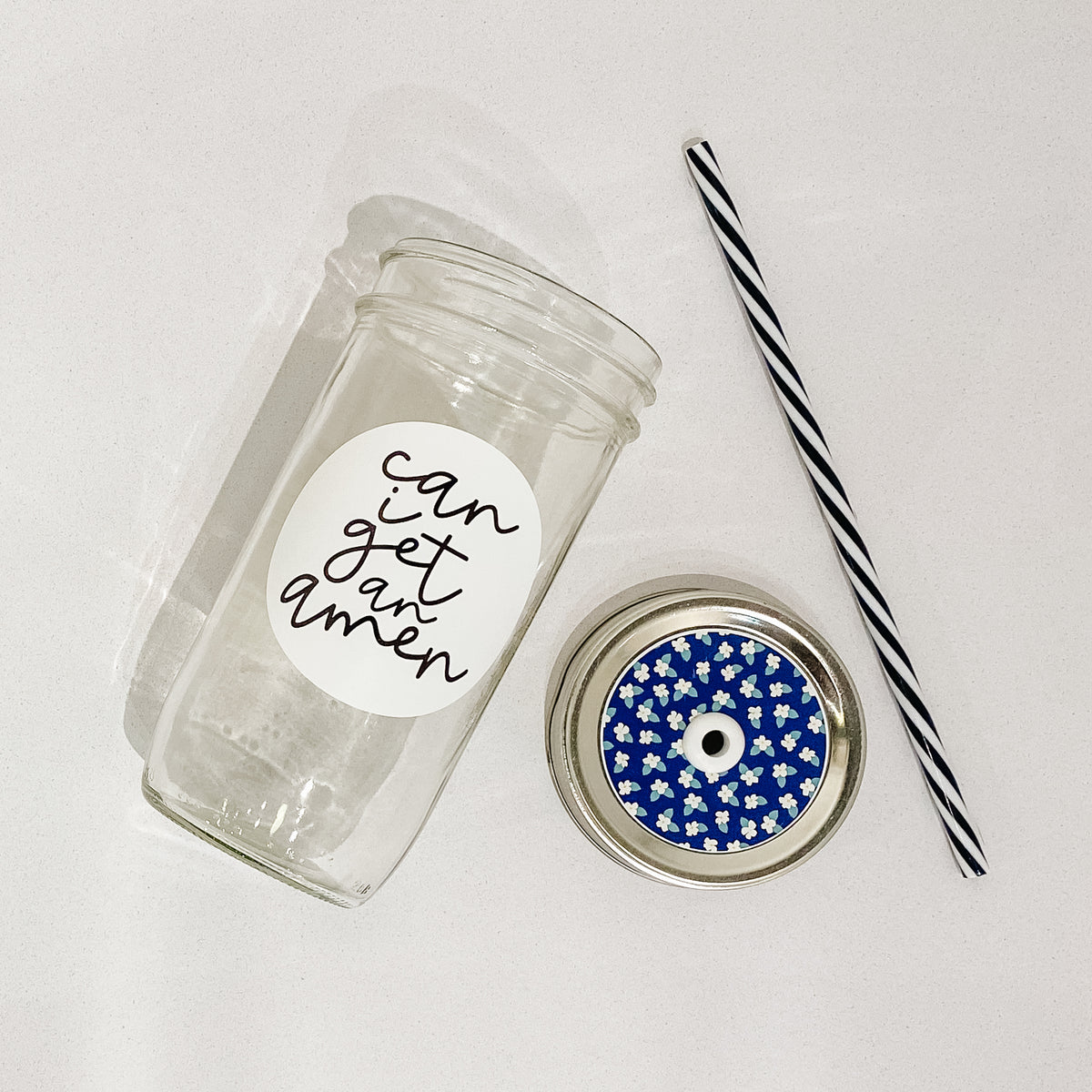 Flat lay of a mason jar tumbler that says “can I get an amen”, a straw lid that is royal blue with white and blue small dainty flowers on it, and a black and white striped straw. 