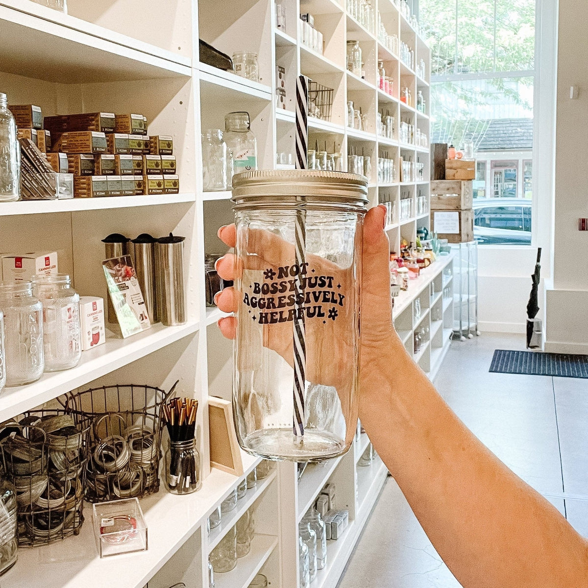 Hand holding a mason jar tumbler that says “not bossy just aggressively helpful” with a small flower at the beginning of the phrase and at the end. All in black. 