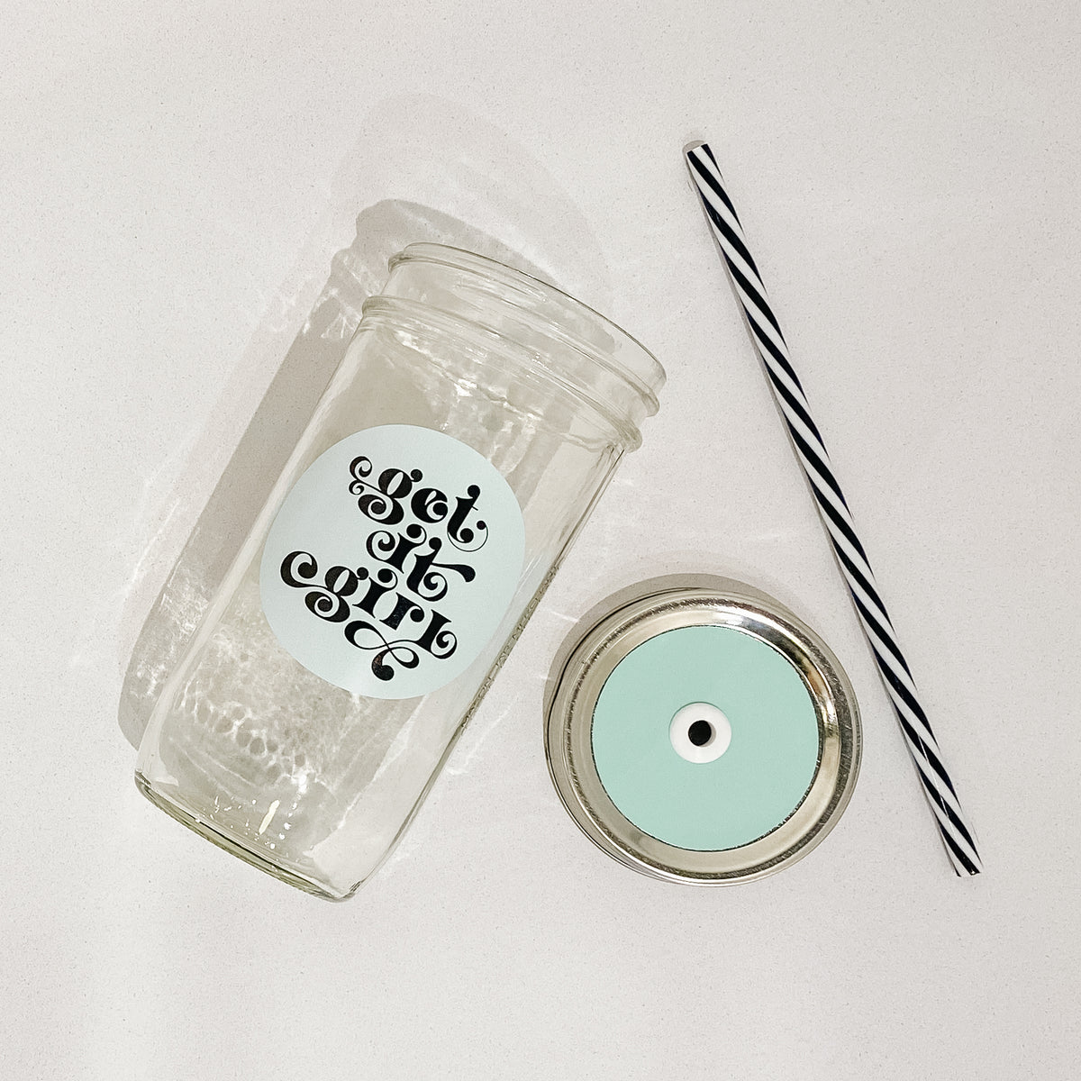 Flat lay of a mason jar tumbler that says “get it girl”, a straw lid that is mint coloured, and a black and white striped straw. 