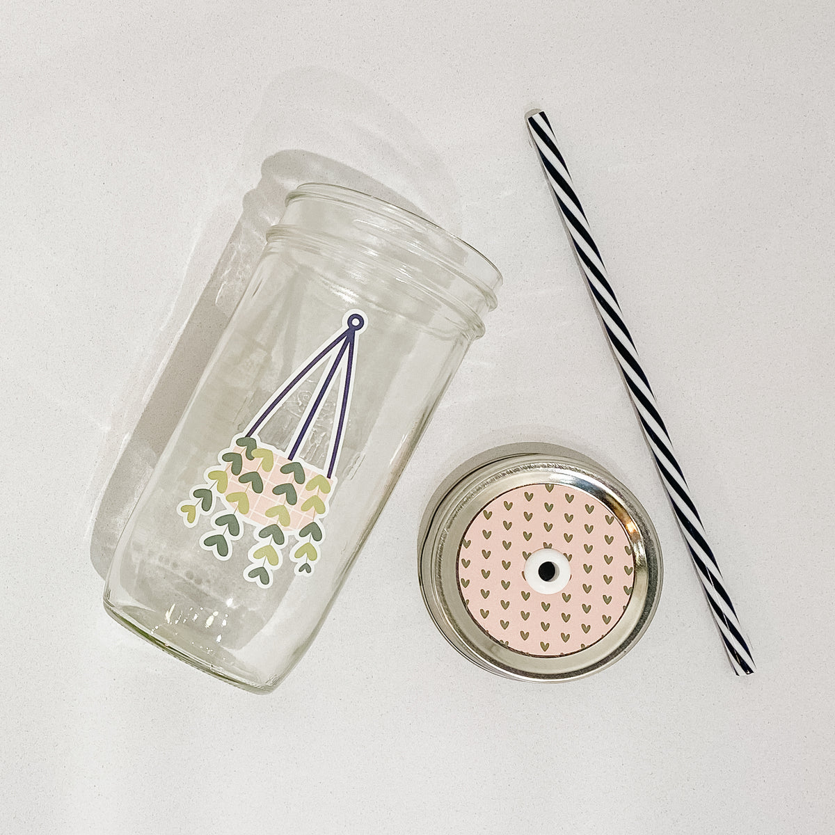 Flat lay of a mason jar tumbler that has a hanging plant decal, a straw lid that has small gold hearts in a repeating pattern on a pink background, and a black and white striped straw. 