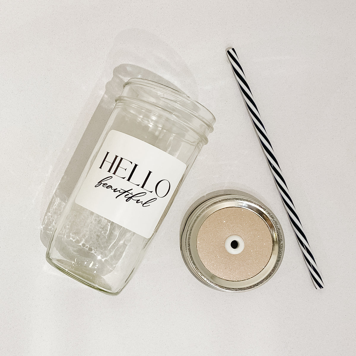 Flat lay of a mason jar tumbler that says “hello beautiful”, a champagne coloured glitter straw lid, and a black and white striped straw