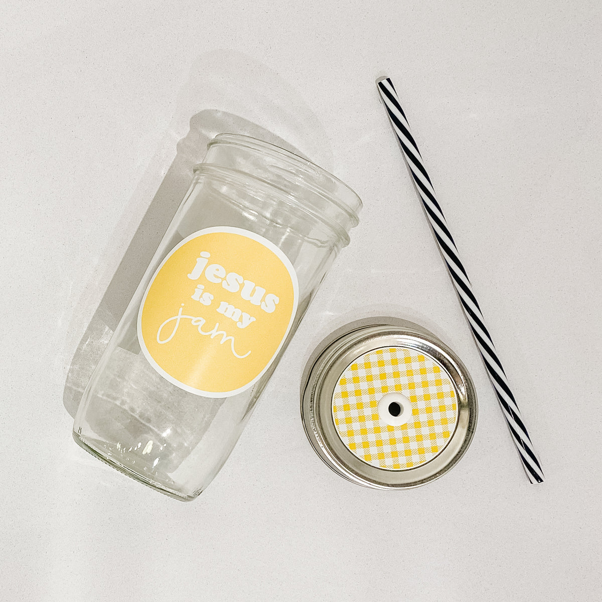 Flat lay of a mason jar tumbler that says “jesus is my jam”, a white and yellow gingham straw lid, and a black and white striped straw. 