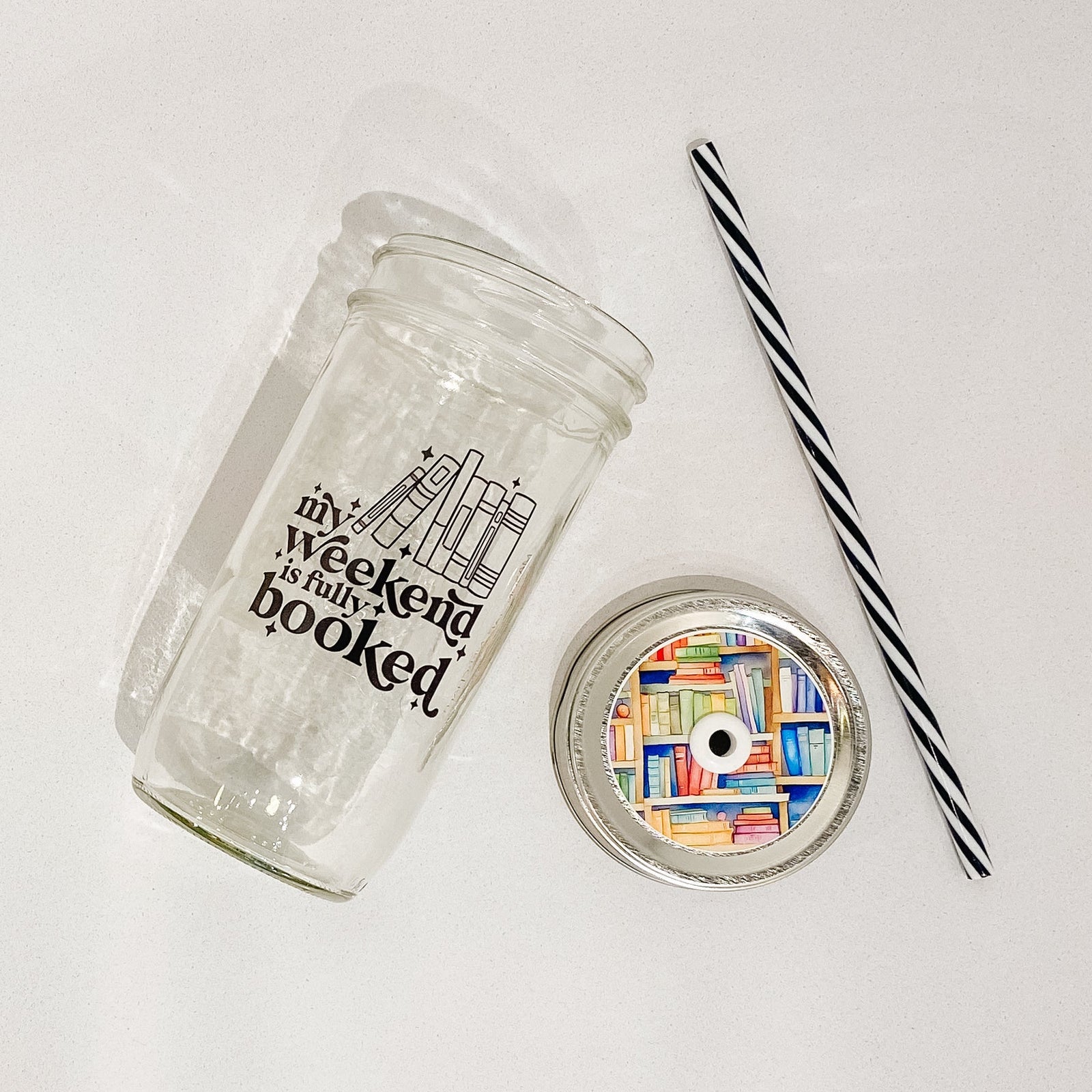 Flat lay of a mason jar tumbler that says “my weekend is fully booked”, a straw lid that is a bookshelf full of multicoloured books, and a black and white striped straw
