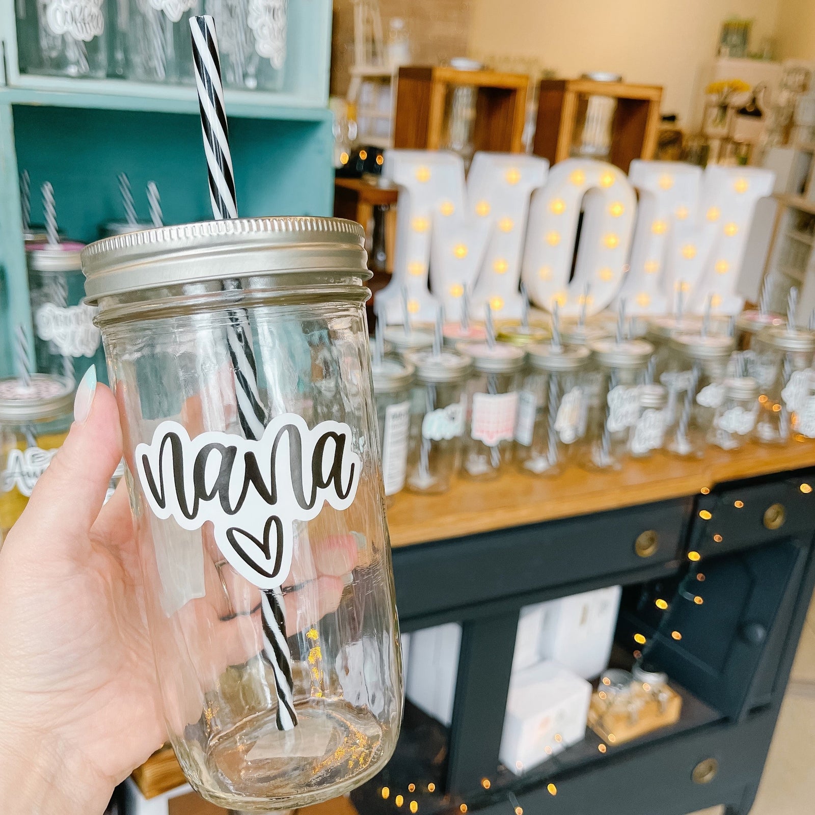 Hand holding a mason jar tumbler that says “nana” with a heart under the text. Text in black script on a white background