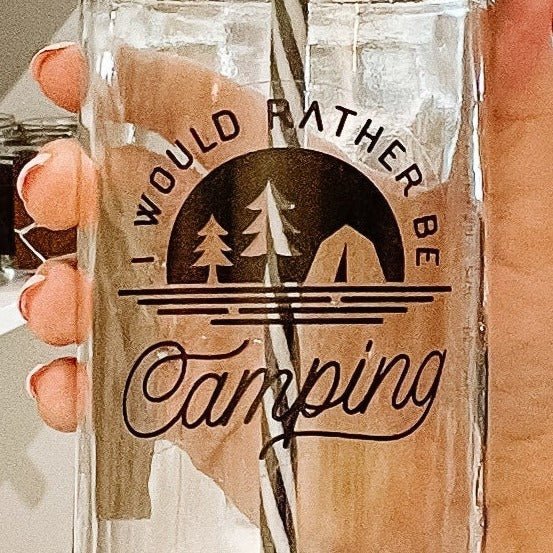 Hand holding a mason jar tumbler that says "I would rather be camping” with clear silhouettes of two trees and a tent on a black background. 