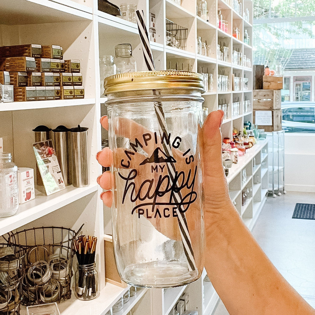 Hand holding a mason jar tumbler that says “camping is my happy place” with a small mountain peak, all in black on a clear background. 