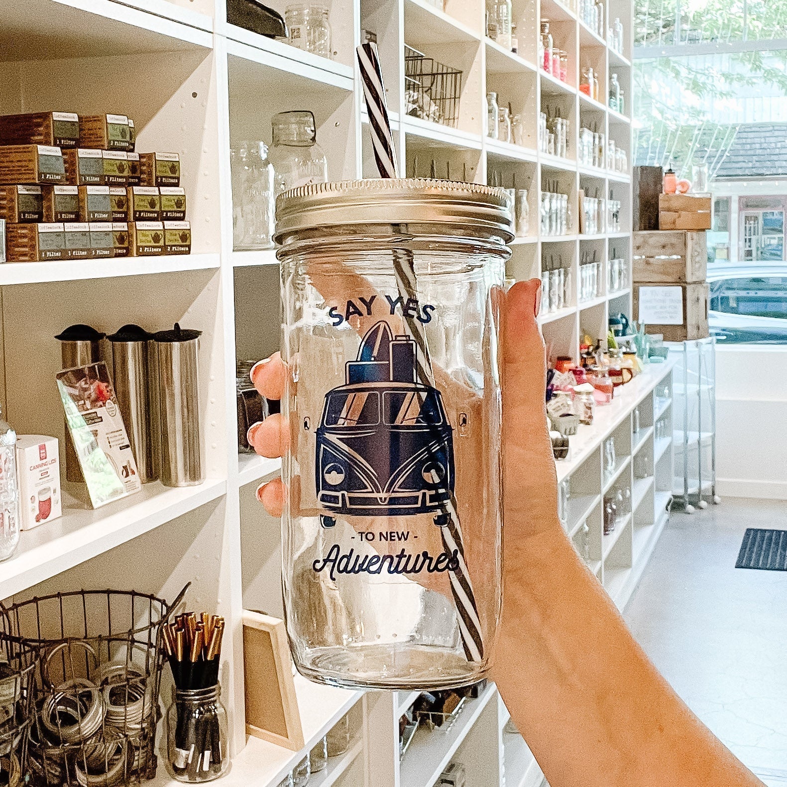 Hand holding a mason jar tumbler that says “say yes to new adventures” with a VW-style van that has suitcases and a surfboard on the roof. All in navy blue on a clear background.