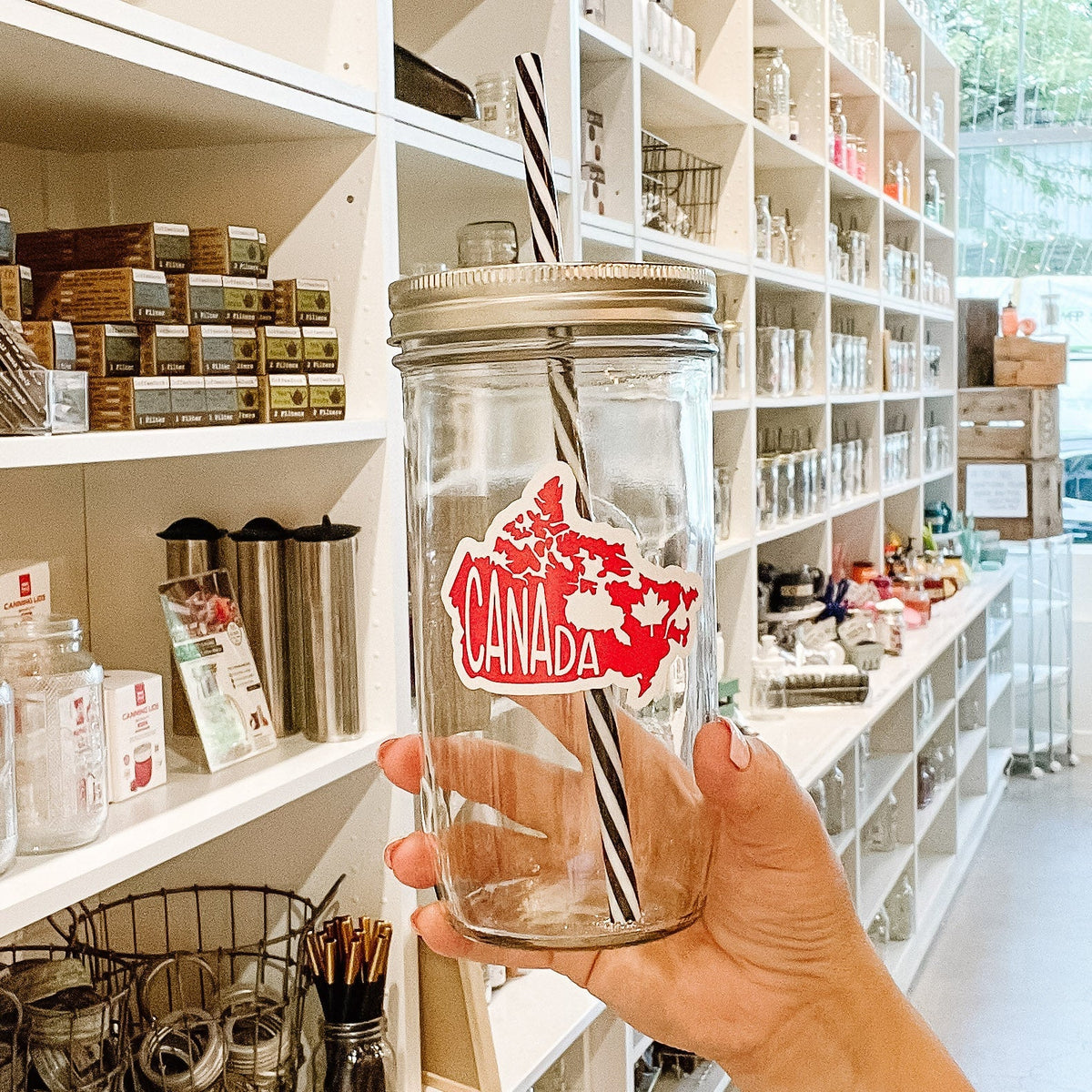 Close up of a hand holding a mason jar tumbler that has an image of Canada filled in red with white lettering saying “Canada”.