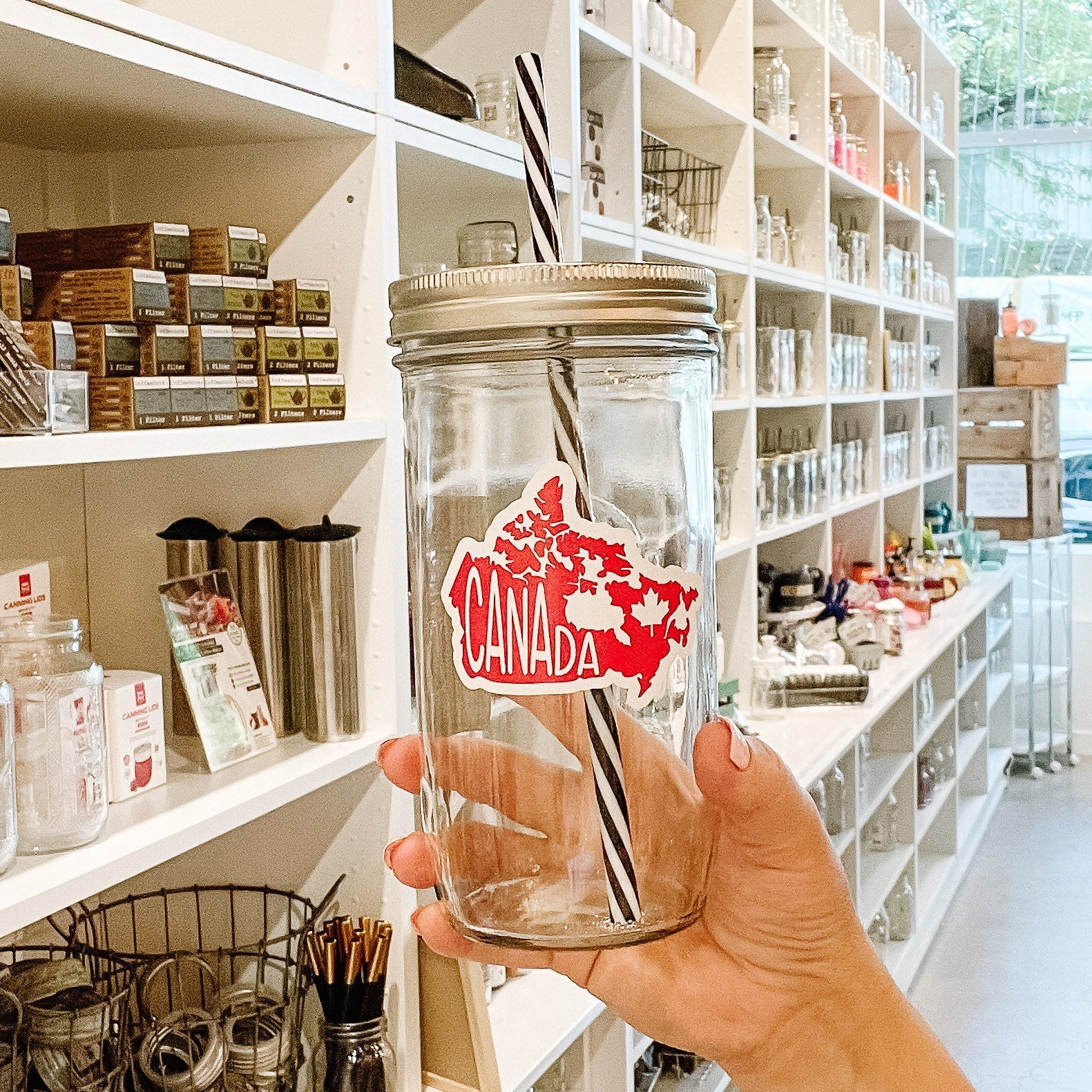Hand holding a mason jar tumbler that has an image of Canada filled in red with white lettering saying “Canada”. 