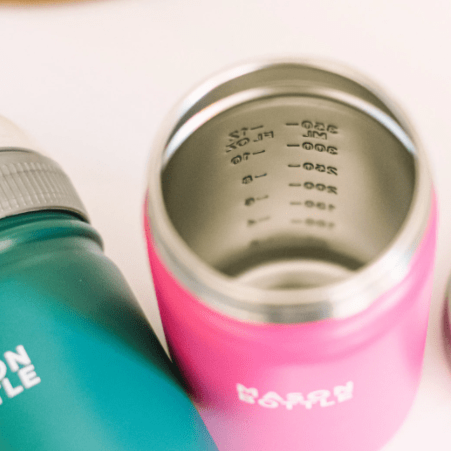 Close up photo of stainless steel mason bottle in pink showing volume markings beside a stainless steel mason bottle in forest green. Photographed against a white countertop.