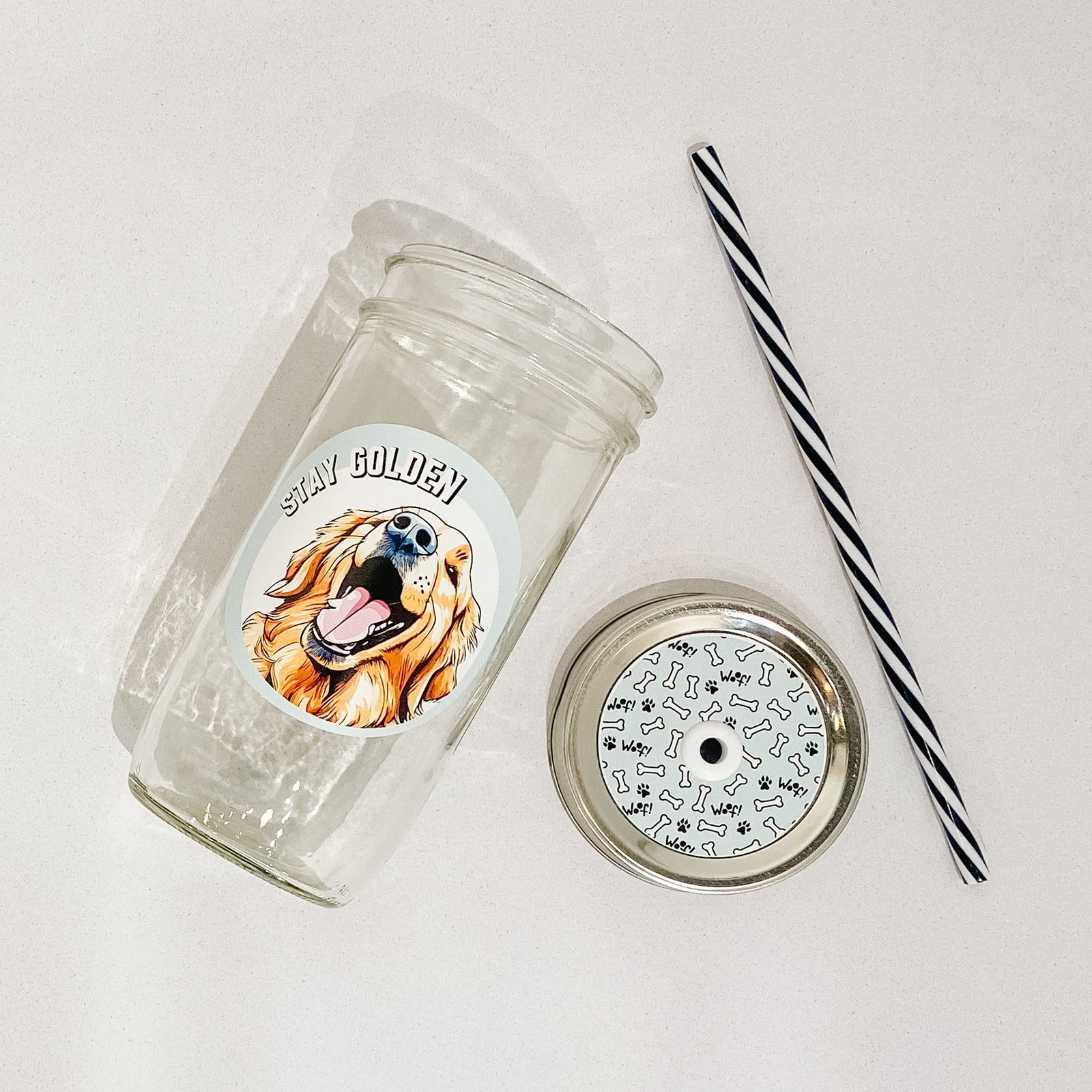 Flat lay of a mason jar tumbler that says "stay golden”, a straw lid with dog bones and paw prints, and a black and white striped straw. 