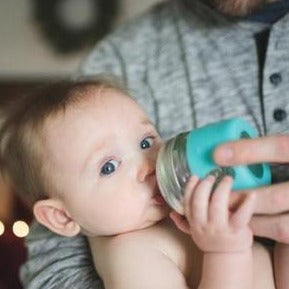 a man holds a baby drinking out of a 8 oz glass reusable mason jar with a teal silicone sleeve and a rubber top lid