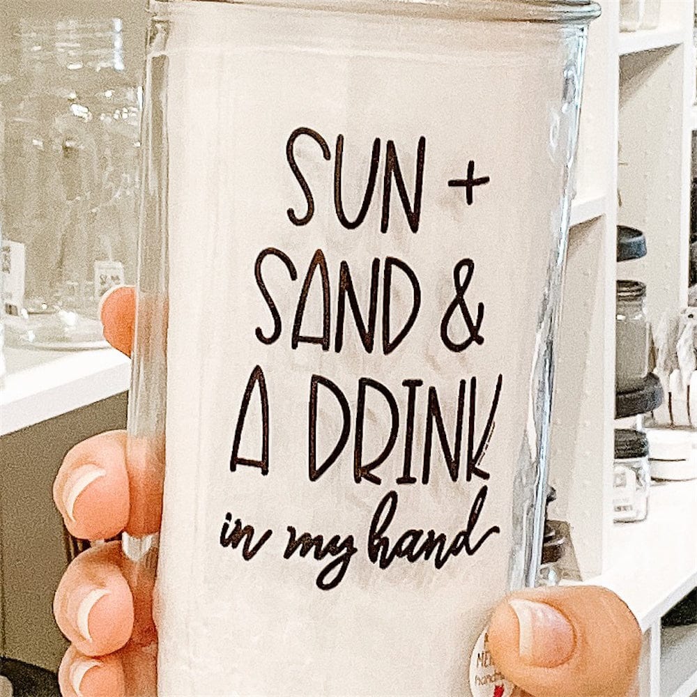 Photo of a mason jar tumblr with a sticker text that says &quot;Sun + Sand &amp; a Drink in my hand&quot;. It has gold lid with black and white straw.A photo of a mason jar tumbler with a sticker text that says &quot;Sun + Sand &amp; A Drink in my hand&quot;