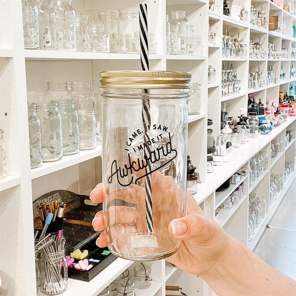 Photo of Mason Jar Tumbler with a gold lid and black and white spiral pattern straw. Mason Jar has a vinyl decal. Photographed against shelves with different mason jars.