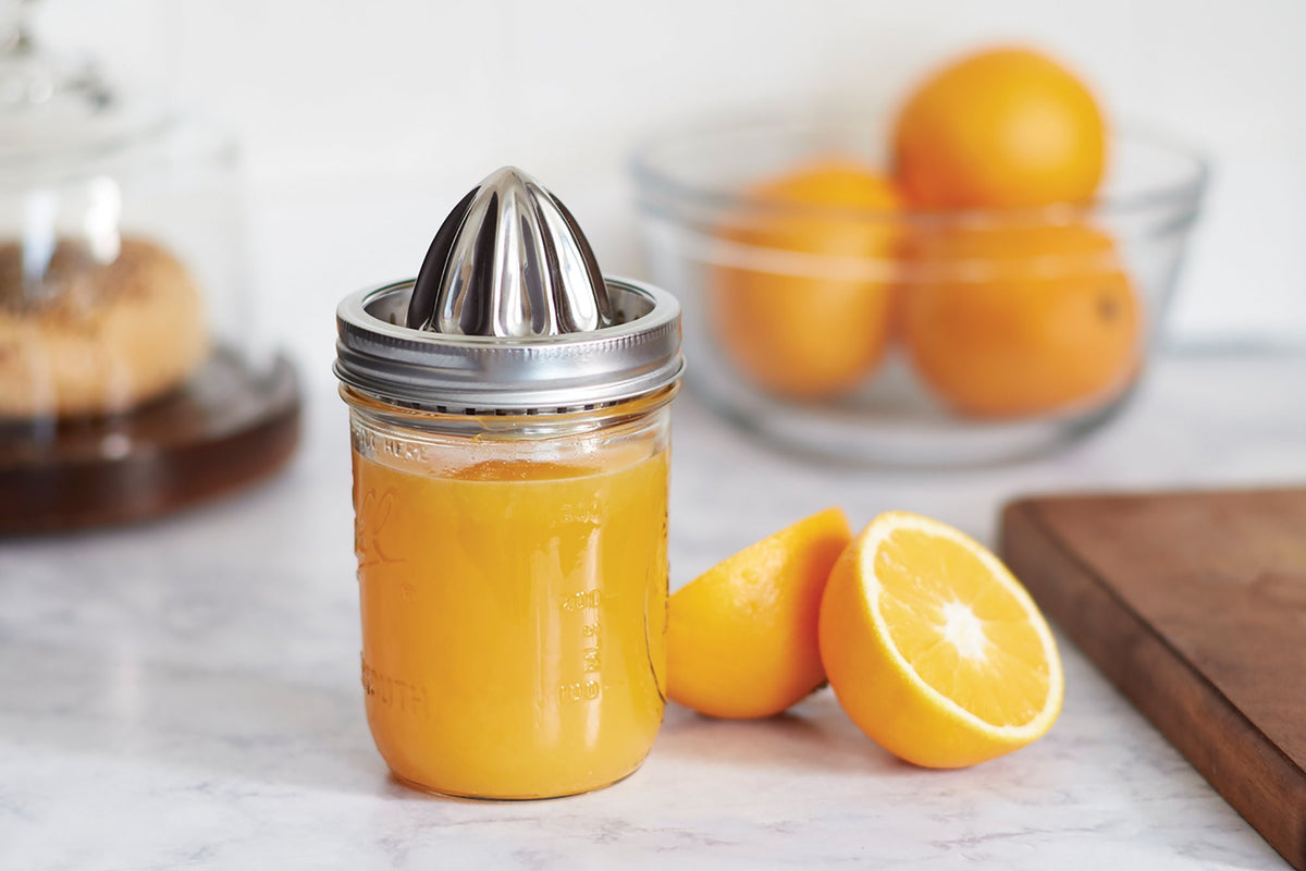 Stainless steel citrus juicer fitted onto a pint size mason jar filled with orange juice, with oranges in the background