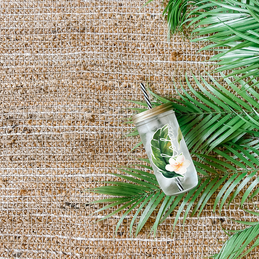 Tumbler with water and a sticker that reads "Aloha" tropical leaf and white hibiscus flower. Photographed as a flat lay in a weave mat and palm leaves.