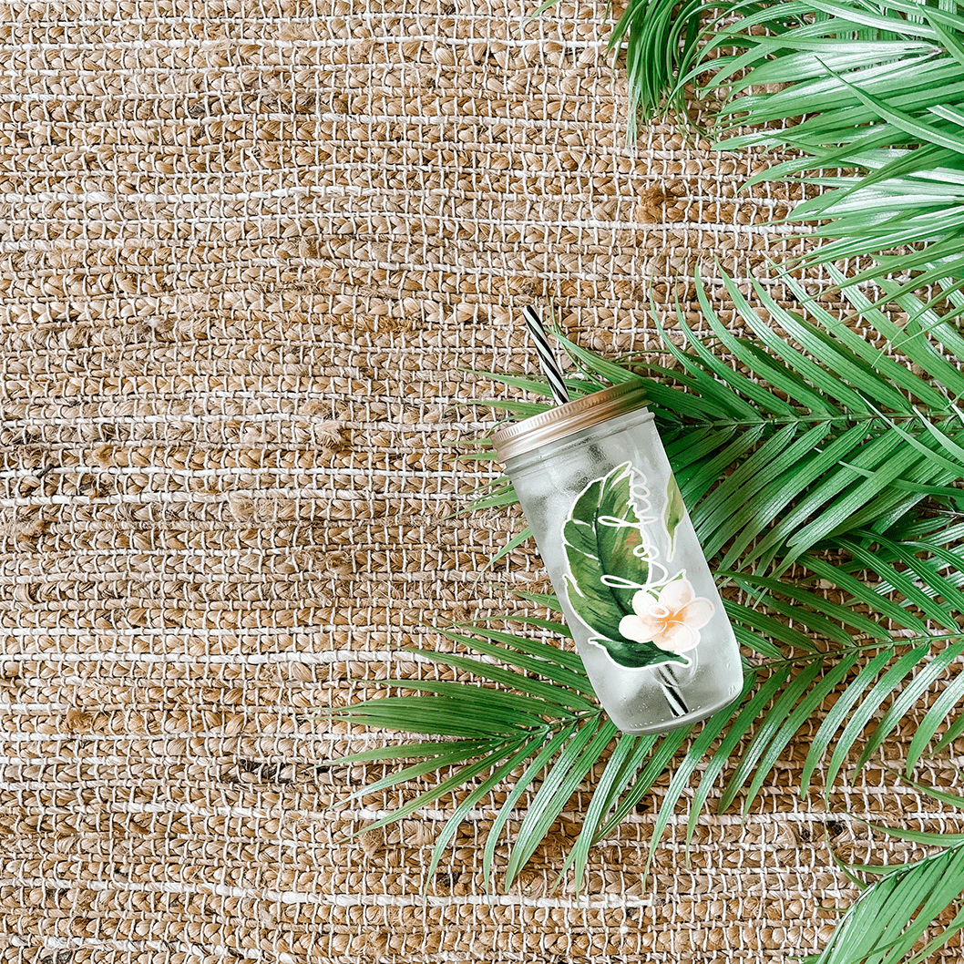 Tumbler with water and a sticker that reads "Aloha" tropical leaf and white hibiscus flower. Photographed as a flat lay in a weave mat and palm leaves.